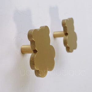 May include: Two gold-colored bear-shaped hooks mounted on a white wall. The hooks have a simple, rounded design and are perfect for hanging items in a nursery or child's room.