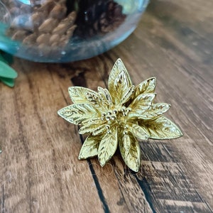 Puede incluir: Un anillo de servilleta de metal dorado en forma de flor con un diseño detallado. El anillo está sobre una superficie de madera.