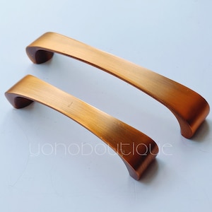 Puede incluir: Dos tiradores de armario de cobre cepillado, uno más grande que el otro, con forma curva.