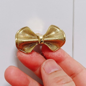 Op de afbeelding: Een goudkleurige kastknop in de vorm van een strik. De knop heeft een getextureerd oppervlak en wordt door een hand tegen een witte achtergrond gehouden. Het strikdesign is gecentreerd met een klein, rechthoekig detail.