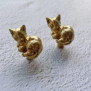 May include: Two gold-colored metal cat figurines, each with a curled-up sleeping pose. The cats are detailed with realistic features and are perfect for adding a touch of whimsy to any space.