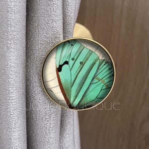 May include: A circular, gold-coloured curtain tieback featuring a teal and black butterfly wing design. The tieback is attached to a grey curtain. The design shows detailed butterfly wing patterns.
