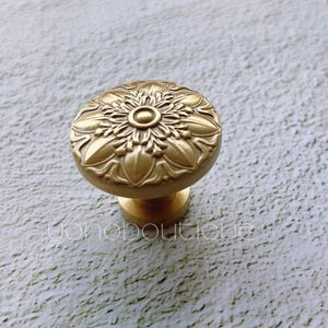 Puede incluir: Un pomo de armario dorado con un diseño floral. El pomo tiene una base redonda y un patrón floral en relieve.