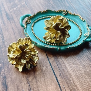 以下が含まれることがあります： ビーズの縁取りが施されたターコイズブルーのトレイの上に、金色の花の形をした装飾品が2つ置かれています。トレイは縁に装飾的な装飾が施されています。