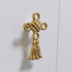 Könnte beinhalten: Goldfarbener Schrankknopf mit Quasten-Design. Der Knopf ist knotenförmig mit einer dekorativen Quaste, die unten hängt.