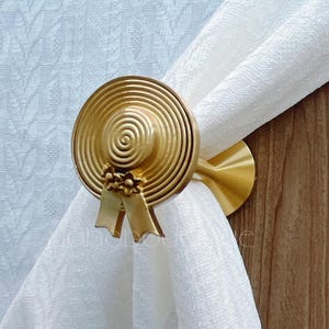 Peut inclure: Un embrasse-rideau doré en forme de chapeau, avec un motif en spirale et un détail de nœud. Il maintient un rideau blanc contre une surface en bois. L'embrasse-rideau en forme de chapeau ajoute une touche décorative.