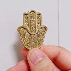 Puede incluir: Un amuleto de mano de hamsa de color dorado con una superficie texturizada.