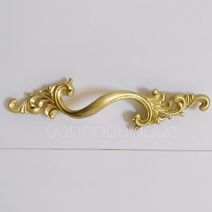 May include: Gold ornate drawer pull with a decorative, swirling design. The pull is shaped like an 'S' and has a smooth, polished finish.