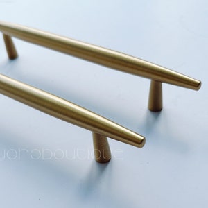 May include: Two gold-toned cabinet pulls with a long, slender design. The pulls have a pointed end and a slightly flared base.