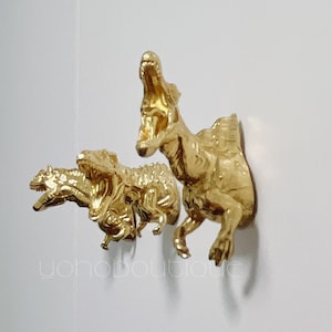 Peut inclure: Trois crochets muraux en forme de dinosaure dorés. Les dinosaures sont alignés, le plus grand à l'arrière et le plus petit à l'avant. Les dinosaures sont tous tournés vers l'avant, la gueule ouverte.