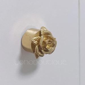 Op de afbeelding: Een goudkleurige kastknop met een rozenontwerp. De roos staat in volle bloei met gedetailleerde bloemblaadjes en bladeren. De knop is op een wit oppervlak gemonteerd. De tekst "yohoboutique" is zichtbaar.
