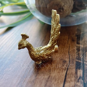 May include: A small, gold-coloured peacock figurine. The bird is detailed with a textured body and a fanned tail. The figurine is set on a wooden surface, with a glass container and greenery in the background.