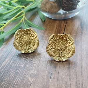 Peut inclure: Une paire de boucles d'oreilles en forme de fleur dorées. Les boucles d'oreilles sont en métal et ont une surface texturée.