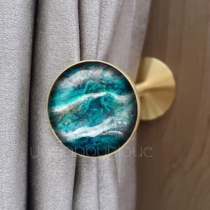 May include: A round curtain tieback with a brass finish. The tieback has a circular design with a teal, white, and blue abstract pattern. The tieback is attached to a grey curtain.