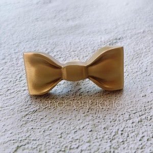 Puede incluir: Un tirador de armario dorado en forma de lazo. El herraje tiene un acabado mate y un diseño clásico de pajarita. El producto es de metal y es adecuado para muebles o aplicaciones decorativas. El texto "jonoboutique" es visible.