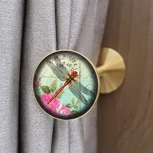 May include: A brass curtain tieback with a circular decorative element. The design features a red and black dragonfly on a vintage floral and script background. The tieback is attached to a grey curtain.