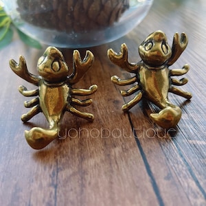 以下が含まれることがあります： 2つのアンティークゴールドカラーの金属製サソリの置物。各サソリは、ハサミ、脚、湾曲した尾を持つ詳細なデザインです。置物は木製の表面にあります。「yohoboutique」というテキストが表示されます。