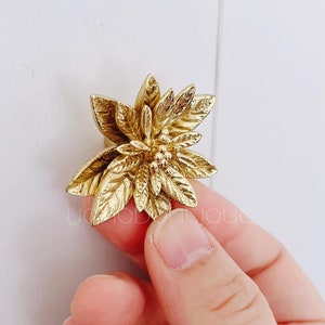 Puede incluir: Un pomo de armario dorado con forma de flor. El pomo presenta diseños detallados de hojas y pétalos, creando un efecto tridimensional. El pomo es sostenido por una mano sobre un fondo blanco.