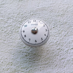 Puede incluir: Pomo de cerámica blanco con números negros del 1 al 12 y la palabra "PARIS" en el centro. El pomo tiene un centro de metal plateado.