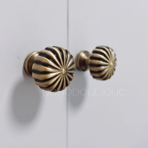 Puede incluir: Dos pomos de armario de latón con un diseño de sol. Los pomos son de color latón oscuro con un centro negro.