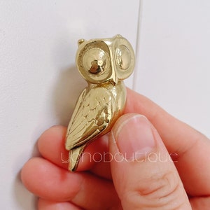 May include: A gold owl figurine with detailed wings and large eyes. The owl is perched on a finger.