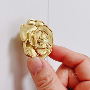 Może przedstawiać: Złoty metalowy gałka w kształcie kwiatu z detalicznym wzorem. Gałka jest trzymana przez rękę.