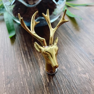 Op de afbeelding: Een gouden metalen flesstopper in de vorm van een hertenkop. De hertenkop heeft een gedetailleerd ontwerp met geweien en een realistisch gezicht.