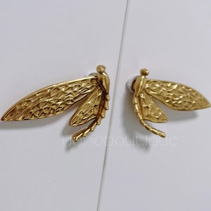 May include: Two gold-colored dragonfly cabinet pulls. Each pull features detailed wings and body, mounted on a white surface. The pulls are designed to be mounted on a cabinet or drawer. The text "yonoboutique" is visible.