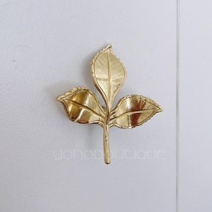 Könnte beinhalten: Eine goldfarbene, blattförmige Brosche mit drei Blättern und einem Stiel. Die Brosche hat detaillierte Adern auf den Blättern und ist vor einem weißen Hintergrund platziert. Der Text "yonoboutique" befindet sich unten.