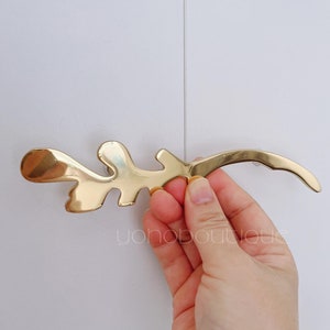 Może przedstawiać: Złoty uchwyt do drzwi szafki w kształcie liścia. Uchwyt ma kształt gałęzi z kilkoma liśćmi.