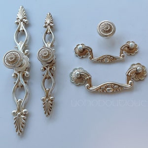 May include: A set of five silver-toned decorative cabinet knobs and pulls. The knobs have a floral design and the pulls have a floral design with a curved shape.