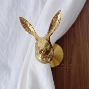 May include: A gold-coloured metal curtain tieback in the shape of a rabbit's head. The rabbit has long ears and detailed facial features. The tieback is mounted on a round base and holds a white curtain.