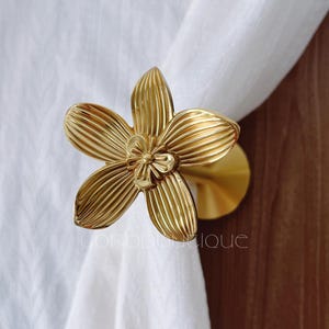 以下が含まれることがあります： 金色の花の形をしたカーテンタッセル。金属製のタッセルは、詳細な花の模様が特徴です。カーテンタッセルは白いカーテンを保持しています。