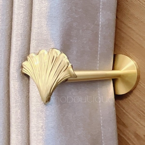 May include: A gold-colored curtain tieback with a fan-shaped design. The tieback is attached to a curtain and has a round base. The curtain is a light beige color, and the background is a wooden surface.