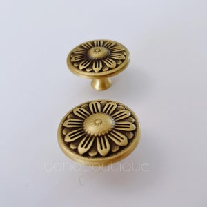 May include: Two gold-toned metal cabinet knobs with a floral design. The knobs have a round base and a raised floral design in the center.