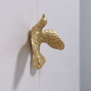 Pode incluir: Figurinha de beija-flor de metal dourado com as asas abertas. A figurinha está montada numa parede branca.