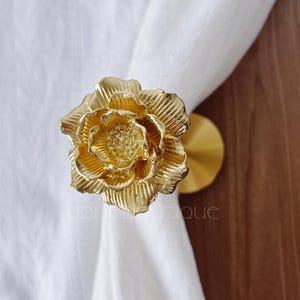 May include: A gold-coloured, flower-shaped curtain tieback. The tieback is made of metal and has a detailed, textured design. The flower is attached to a round, gold-coloured base.