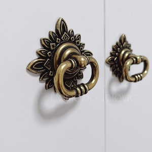 Puede incluir: Dos tiradores de puerta de armario de color dorado adornados con un diseño floral. El tirador más grande tiene una manija en forma de anillo, mientras que el tirador más pequeño tiene una manija de bucle más tradicional.