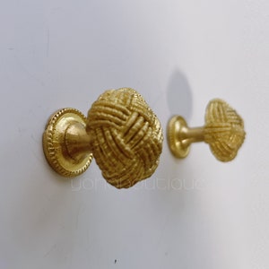May include: Two gold-toned cabinet knobs with a rope-like design. The knobs are mounted on a white wall.