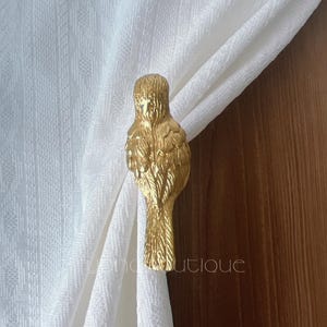 May include: A gold-coloured bird-shaped curtain tieback. The bird has detailed feather carvings and is holding a white curtain. The background includes a wooden surface and a white patterned curtain.