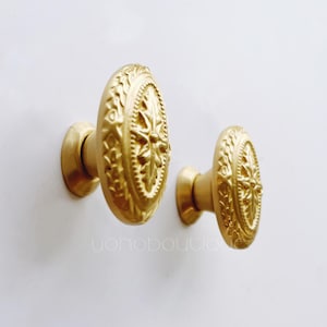 Könnte beinhalten: Zwei goldfarbene Schrankknöpfe mit einem verzierten Design. Die Knöpfe sind oval geformt und haben ein erhabenes, detailliertes Muster.