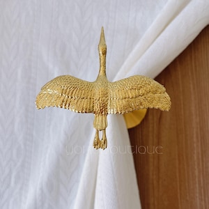 May include: A gold-colored metal curtain tieback in the shape of a crane with outstretched wings. The bird is attached to a round base, holding white fabric. The tieback is a decorative element for window treatments.