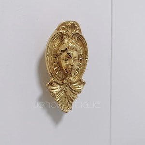 Op de afbeelding: Een gouden metalen kastknop met een gedetailleerd ontwerp met een vrouwengezicht en een decoratieve strik.