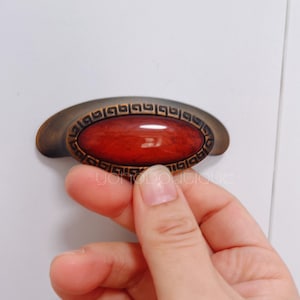 Op de afbeelding: Een ovale kastknop in rood en goud met een decoratieve rand. De knop is gemaakt van metaal en heeft een gladde, gepolijste afwerking.