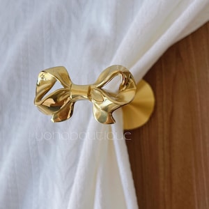 May include: A gold-coloured curtain tieback shaped like a bow. The bow is made of a shiny, metallic material and is attached to a round base. The tieback is holding a white curtain.