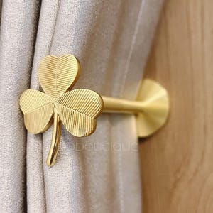 Może przedstawiać: Złoty uchwyt do zasłon w kształcie trójlistnej koniczyny. Uchwyt jest przymocowany do okrągłej podstawy i trzyma beżową zasłonę. Koniczyna ma szczegółowe żyłki liści.