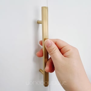 Pode incluir: Uma puxador de armário dourado com forma retangular e bordas ligeiramente arredondadas. O puxador está montado numa superfície branca.