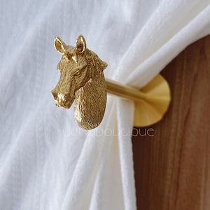 May include: A gold-colored curtain tieback featuring a detailed horse head design. The tieback is holding a white curtain. The horse head is intricately sculpted with a textured surface. The design includes a round base and a rod.