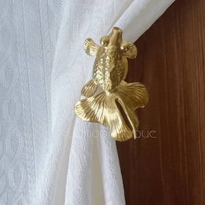 Könnte beinhalten: Ein goldfarbener, fischförmiger Vorhanghalter. Der Halter ist aus Metall gefertigt und hat ein detailliertes Design, einschließlich Flossen und Schuppen. Er hält einen weißen Vorhang.