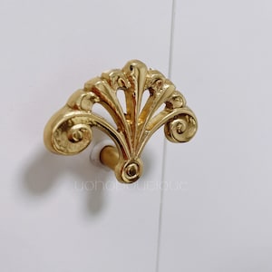 Könnte beinhalten: Ein goldfarbener, dekorativer Schrank-Knopf mit einem fächerförmigen Design. Der Knopf hat ein detailliertes, verziertes Design mit Schnörkeln und Kurven.